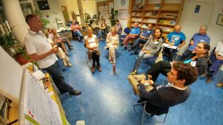 Foto: Genossenschaft für Gemeinwohl Menschen sitzen im Sesselkreis, Mann steht links und redet